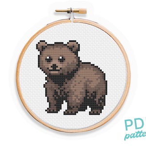 May include: A brown bear cub cross stitch pattern in a wooden embroidery hoop. The pattern is pixelated and features a brown bear cub with a white muzzle and black eyes.