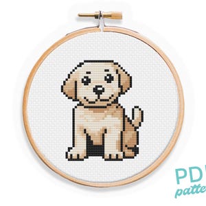 May include: Cross stitch pattern of a cute, cartoon-style, light brown puppy sitting with its tongue out. The pattern is in a wooden embroidery hoop.