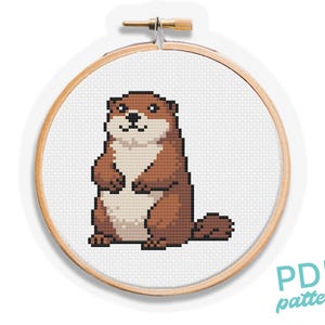 May include: A cross-stitch pattern of a cartoon otter in a wooden embroidery hoop. The otter is brown and beige, with a pixelated design. The text "PDF pattern" is in the lower right corner.