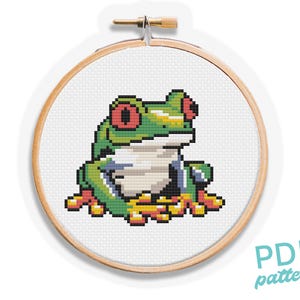 Könnte beinhalten: Ein Kreuzstichmuster einer grünen und gelben Frosch mit roten Augen, der auf einem weißen Hintergrund sitzt. Das Muster befindet sich in einem hölzernen Stickrahmen.