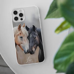 May include: Clear phone case featuring a photograph of two horses. One horse has a light brown coat and a white mane, while the other is black. The word "Liv" is written in cursive at the bottom of the case.