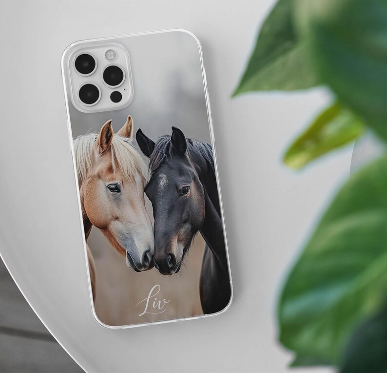 May include: Clear phone case featuring a photograph of two horses. One horse has a light brown coat and a white mane, while the other is black. The word "Liv" is written in cursive at the bottom of the case.