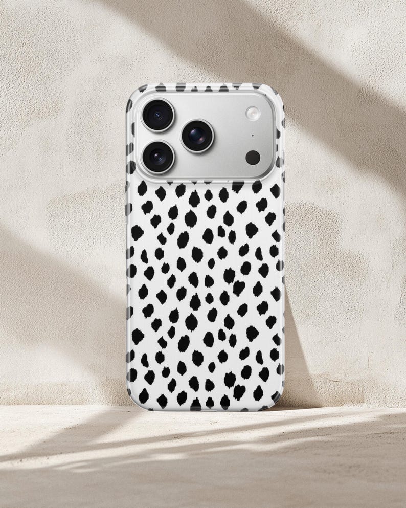 May include: A smartphone case featuring a white background with a repeating pattern of irregular black spots. The case has a clear border and a rectangular cutout at the top for the camera lenses, which are visible as three circular black circles.