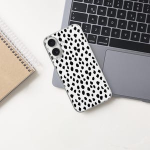 May include: A smartphone case featuring a white background with a repeating pattern of irregular black spots. The case has two circular cutouts for camera lenses on the upper left side and is positioned on a dark gray laptop keyboard.