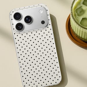 May include: A smartphone case featuring a white background with a repeating pattern of small black polka dots. The case has a clear border and a rectangular cutout at the top left corner to accommodate a triple-lens camera system and a flash.