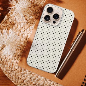May include: A smartphone case featuring a cream-colored background with a repeating pattern of small black polka dots. The case has a rectangular cutout at the top left corner for the camera lenses and is positioned on a brown notebook next to a pen.