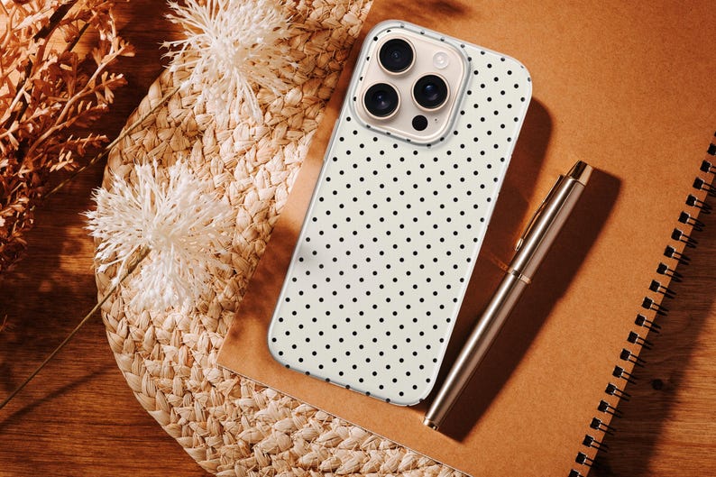 May include: A smartphone case featuring a cream-colored background with a repeating pattern of small black polka dots. The case has a rectangular cutout at the top left corner for the camera lenses and is positioned on a brown notebook next to a pen.