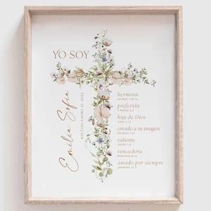 May include: Framed print featuring a floral cross design with the text "YO SOY" and "Emilia Sofia." The cross is adorned with flowers and greenery. Additional text includes "hermosa," "preferida," and other Spanish phrases. The frame is light brown.