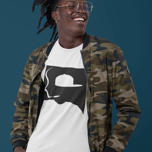 Op de afbeelding: Een persoon draagt een wit T-shirt met een zwart silhouet van een persoon die een hoed draagt. De persoon draagt een camouflagejas en heeft dreadlocks.