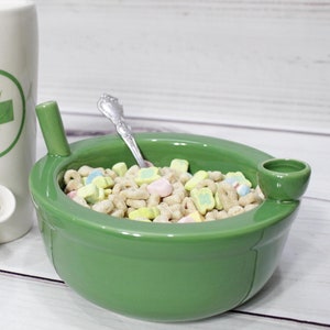 K&ouml;nnte beinhalten: Eine gr&uuml;ne Keramiksch&uuml;ssel mit einem eingebauten Ausguss, gef&uuml;llt mit M&uuml;sli und Marshmallows. Die Sch&uuml;ssel hat einen L&ouml;ffel, der auf dem Rand liegt.