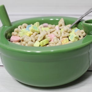 K&ouml;nnte beinhalten: Eine gr&uuml;ne Keramiksch&uuml;ssel mit einem Ausguss und einem Griff, gef&uuml;llt mit Fr&uuml;hst&uuml;cksflocken mit Marshmallows.