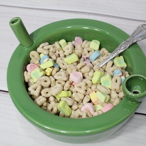 K&ouml;nnte beinhalten: Eine gr&uuml;ne Keramiksch&uuml;ssel mit einem Ausguss auf jeder Seite, gef&uuml;llt mit einem Fr&uuml;hst&uuml;cksm&uuml;sli mit Marshmallows. Ein silberner L&ouml;ffel befindet sich in der Sch&uuml;ssel.
