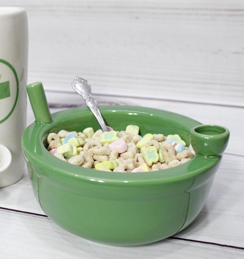 K&ouml;nnte beinhalten: Eine gr&uuml;ne Keramiksch&uuml;ssel mit einem eingebauten Ausguss, gef&uuml;llt mit M&uuml;sli und Marshmallows. Die Sch&uuml;ssel hat einen L&ouml;ffel, der auf dem Rand liegt.
