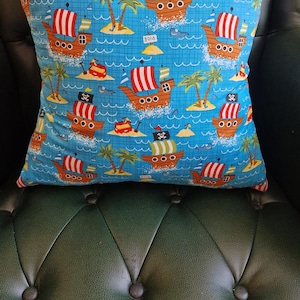 May include: A blue pillow with a repeating pattern of pirate ships, palm trees, and islands. The ships have red and white stripes and a skull and crossbones flag. The background is a light blue with a grid pattern.