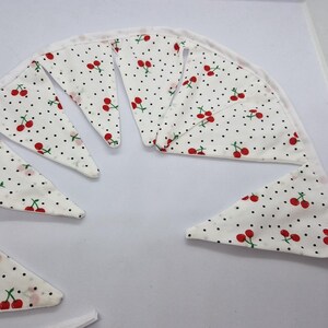 May include: A white fabric bunting featuring a pattern of red cherries and black polka dots. The triangular flags are strung together with a white ribbon, creating a decorative banner. Perfect for parties and celebrations.