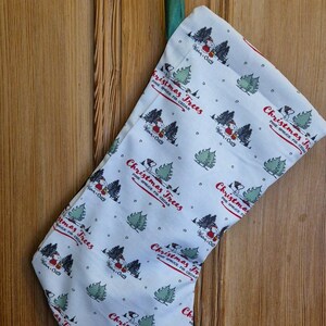 以下が含まれることがあります： 緑色の吊り下げループが付いた白いクリスマスストッキング。ストッキングには、緑のクリスマスツリー、赤と白の家、そして「Christmas Trees」の文字がパターン化されています。木製のドアに吊るされています。