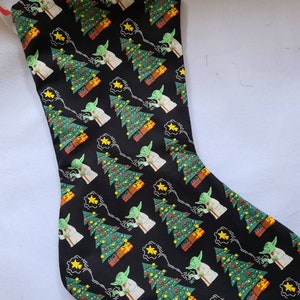 Puede incluir: Calcetín navideño negro con un patrón repetido de un personaje verde, árboles de Navidad y estrellas amarillas. Los árboles están decorados con adornos rojos y amarillos y se encuentran junto a regalos envueltos. Una presilla roja está unida en la parte superior.