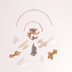 Puede incluir: Un móvil de madera con un elefante de fieltro gris, dos aviones de fieltro marrón y tres nubes de fieltro blanco. El móvil también incluye una esfera de bolas de fieltro marrón, beige y blanco.