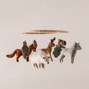 Könnte beinhalten: Ein Baby-Mobile mit Filztierfiguren, darunter ein Fuchs, ein Hirsch, ein Eisbär und ein graues Tier, das an einem Holzring aufgehängt ist. Die Tiere sind in Braun-, Weiß- und Grautönen gehalten, mit grünen Akzenten.