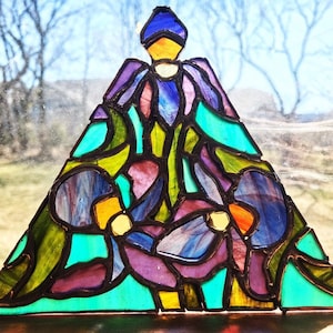 Puede incluir: Una obra de arte en vidriera con forma de ángel. La obra presenta una vibrante mezcla de colores, incluyendo azul, morado, verde, amarillo y naranja, con contornos negros. El ángel está situado sobre un fondo exterior borroso.