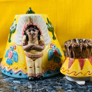 Könnte beinhalten: Eine Teekanne aus Keramik in Form eines indianischen Tipis mit einer Indianerfigur, die im Eingang steht. Die Teekanne ist mit bunten Vögeln und geometrischen Mustern verziert. Der Deckel ist separat und hat die Form eines Kopfschmucks.