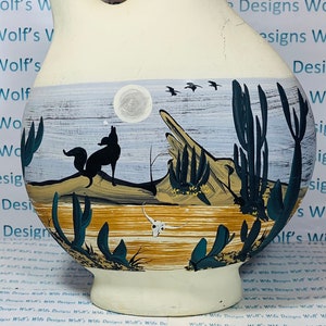 Könnte beinhalten: Ein weißer Keramik Krug mit einer Wüstenlandschaft, die auf die Vorderseite gemalt ist. Die Szene zeigt einen Kojoten, Kakteen und einen Vollmond. Der Krug hat eine einzigartige Form mit einer großen Öffnung oben und einem schmalen Ausguss.