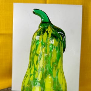 Könnte beinhalten: Eine dekorative Glas-Birnen-Skulptur. Die Birne ist hauptsächlich grün mit gelben Akzenten und einem grünen Stiel. Das Glas hat ein gesprenkeltes Muster. Die Skulptur steht auf einer dunkelgrauen Oberfläche.