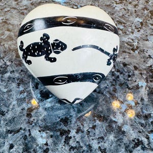 May include: Heart-shaped, off-white decorative object with black horizontal bands and stylized animal figures. The bands have curved, eye-like designs. The animal figures are black with white dot patterns. The object is on a reflective surface.