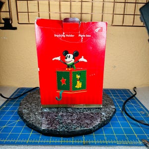 May include: A red Christmas stocking holder featuring Mickey Mouse. The box has the words "Stocking Holder" and "Parte bas" printed on it. The holder has a green star and a picture of Pluto on it.