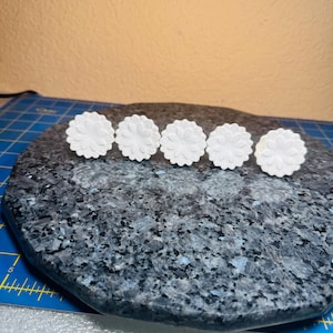 May include: Five white, flower-shaped decorative objects are arranged in a row on a dark, speckled surface. Each object has a detailed, layered petal design. The background is a solid, light-colored wall.