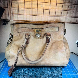 May include: A tan leather handbag with a gold-tone lock and chain strap. The bag has two handles and a decorative leather tag. The Michael Kors logo is visible on the lock. The bag is sitting on a blue surface.