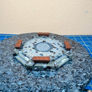 May include: A miniature model of a stone-paved circular plaza with four wooden benches. The plaza is surrounded by a stone wall and sits on a dark, speckled base. The model is likely for a tabletop game or diorama.