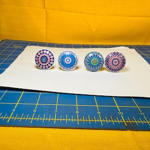 May include: Four decorative cabinet knobs with colorful mandala designs. The knobs feature pink, blue, green, and purple patterns. They are arranged in a row on a white surface, perfect for adding a unique touch to furniture.