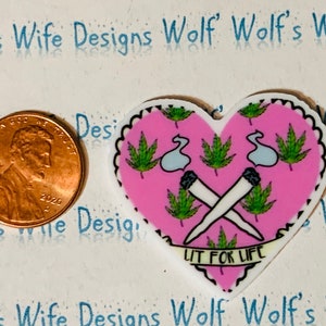 May include: Pink heart-shaped resin charm with a white outline. The charm features a graphic of two crossed marijuana joints with green marijuana leaves and the text "Lit for Life".