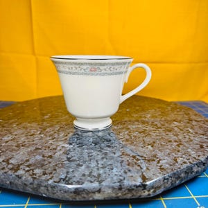 May include: A white teacup with a decorative floral and vine pattern around the rim. The cup has a curved handle and sits on a small pedestal base. The rim is trimmed with a silver band. The background is a solid yellow.
