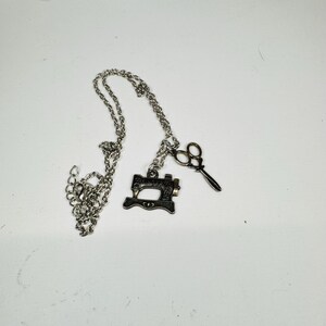May include: A silver-toned metal necklace featuring two charms: a miniature sewing machine and a pair of scissors. The chain is a standard link style. The sewing machine charm has the word SINGER clearly visible on its side in raised lettering.
