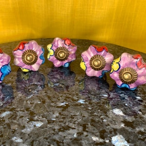 Op de afbeelding: Vijf keramische bloemvormige ladegrepen met een paarse basis en kleurrijke bloemenmotieven. Elke knop heeft een gouden midden.