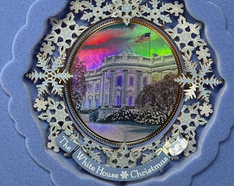 May include: A white and silver Christmas ornament featuring a depiction of the White House with a snowy background and a colorful aurora borealis in the sky. The ornament is surrounded by snowflakes and has the text "The White House Christmas" on the bottom.