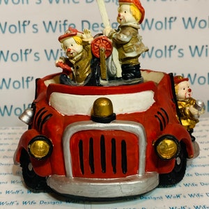 Puede incluir: Una figurita de camión de bomberos rojo con tres payasos montados en la parte superior. Los payasos llevan sombreros rojos y amarillos, chaquetas marrones y pantalones blancos. El camión de bomberos tiene una cabina blanca y una parrilla negra.