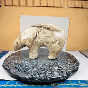 Puede incluir: Una escultura de oso de piedra tallada con un cuerpo color crema y vetas marrones. El oso se encuentra sobre una base de piedra octogonal gris oscuro. La espalda del oso presenta un diseño circular con líneas grabadas.