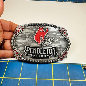 Peut inclure: Une boucle de ceinture ovale en métal argenté avec une texture grain de bois, une silhouette de cheval cabré rouge et le texte "Let'er Buck" et "PENDLETON WHISKY". La bordure est perlée avec des accents rouges, mesurant environ 10 cm de large.