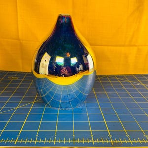 May include: A decorative vase with a glossy, reflective surface. The top is dark blue, transitioning to yellow, and then to a blue grid pattern at the base. The vase is on a blue grid surface with a yellow background.