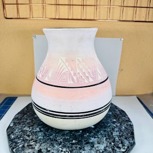Może przedstawiać: Ręcznie wykonany ceramiczny wazon z białą górą, przechodzący w różowo-biały geometryczny wzór i czarny pasek. Wazon stoi na ciemnej, nakrapianej, sześciokątnej powierzchni. Ozdobna ceramika.