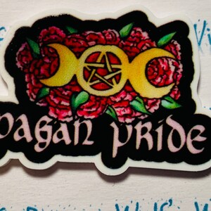 Puede incluir: Una pegatina en blanco y negro con un diseño floral rosa y verde. El centro de la pegatina presenta un símbolo de triple luna dorada con un pentagrama en su interior. El texto "pagan pride" está escrito en una fuente estilizada debajo del símbolo.