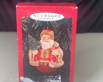 May include: A red and gold Christmas ornament box featuring a Santa Claus holding a small Christmas tree. The box has the text "Keepsake Ornament" and "Special Edition" printed on it.