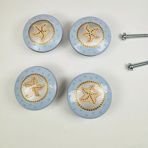 May include: Four round cabinet knobs with a light blue base and small teal polka dots. Each knob features a central beige starfish design surrounded by a ring of gold dots. Two silver metal screws are positioned to the right of the knobs.