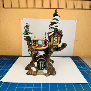 May include: A decorative tree-shaped figurine featuring a brown trunk base with a small house entrance, topped by a platform with two figures, a small cabin with a window, and two snow-dusted evergreen trees decorated with colorful lights.