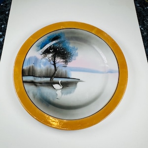 May include: A decorative plate with a landscape scene featuring a tree, a swan, and a body of water. The plate has a gold-coloured rim and a white interior. The artwork includes shades of blue, black, and pink.