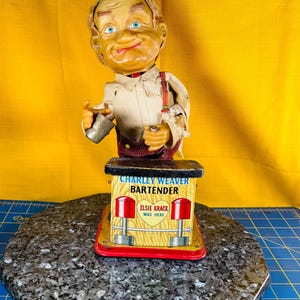 Könnte beinhalten: Ein Vintage-Spielzeug von Charley Weaver Bartender. Das Spielzeug zeigt ein lächelndes Gesicht mit blauen Augen, das ein weißes Hemd, rote Hosenträger und eine braune Perücke trägt. Die Basis trägt den Text "Charley Weaver Bartender" und "Elsie Krack Was Here."