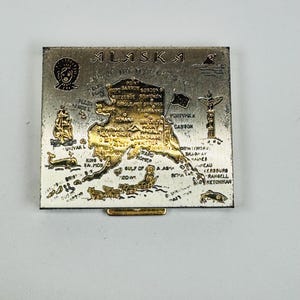 May include: A square metal souvenir compact mirror featuring a gold-toned map of Alaska on a silver background. The map includes engraved text labels for cities and landmarks, along with illustrations of a totem pole, a whale, and a dog sled.
